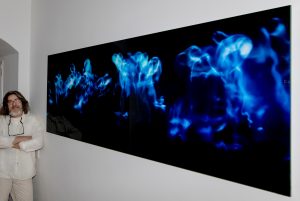 Ein Foto des Fotokünstlers Ralf Lindenau neben seinem aufgehangenen, großformatigen Werk in Blautönen stehend, im Auftrag der Galerie Ahlemann.