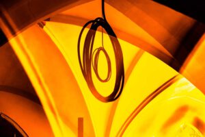 Galerie Ahlemann zeigt ein abstraktes Foto von Ingrid Pohl aus der Konzeptreihe "Zwischen Zeiten" auf dem in orangen und gelben Farbtönen ein eingerolltes Elektrokabel an der Decke eines Kirchengewölbes hängt.