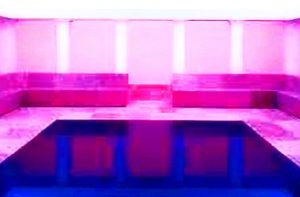 Galerie Ahlemann zeigt ein abstraktes Foto des Fotokünstlers Jochen Cerny in überwiegend rosa und blauen Farbtönen. Das Foto zeigt den Blick in einen rechteckigen Raum , dessen sichtbare Ausenwände mit einer Sitzbank ausgestattes ist, die nur kurz mittig an der Rückwand unterbrochen wird.