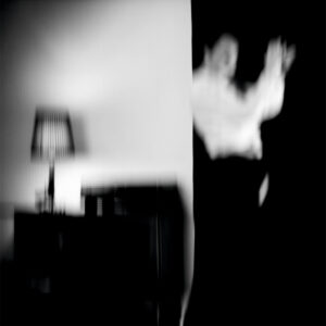 Galerie Ahlemann zeigt ein abstraktes schwarz-weiß Foto von Mick Schäfer aus der Reihe "feminin move" auf dem im rechten Drittel ein Tänzer vor schwarzem Hintergrund zu sehen ist, während auf den linken zwei Drittel eine Kommode mit Lampe vor einer weißen Wand steht.