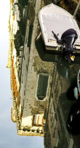 Galerie Ahlemann zeigt ein Foto des Fotokünstlers Mick Schäfer aus der Serie "Reflecting Venice" auf dem ein abgestelltes Motorboot auf auf dem Wasser zu sehen ist, in dessen Oberfläche sich ein mehrstöckiges Haus spiegelt.